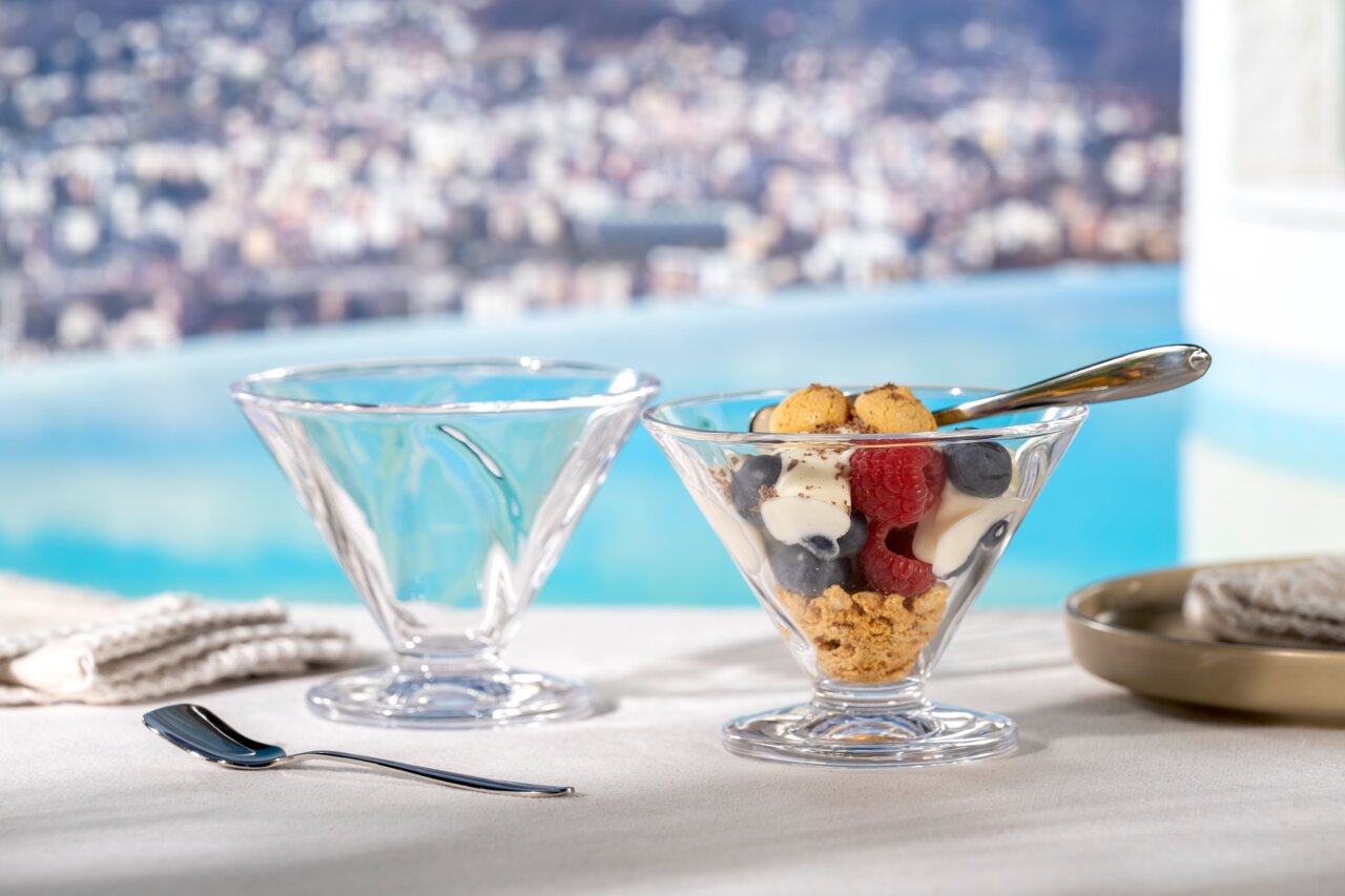 Zwei elegante Glasschalen auf Fuß, eine gefüllt mit Dessert und Beeren, vor einem verschwommenen Hintergrund mit Pool und Stadtansicht, aus seitlicher Perspektive.