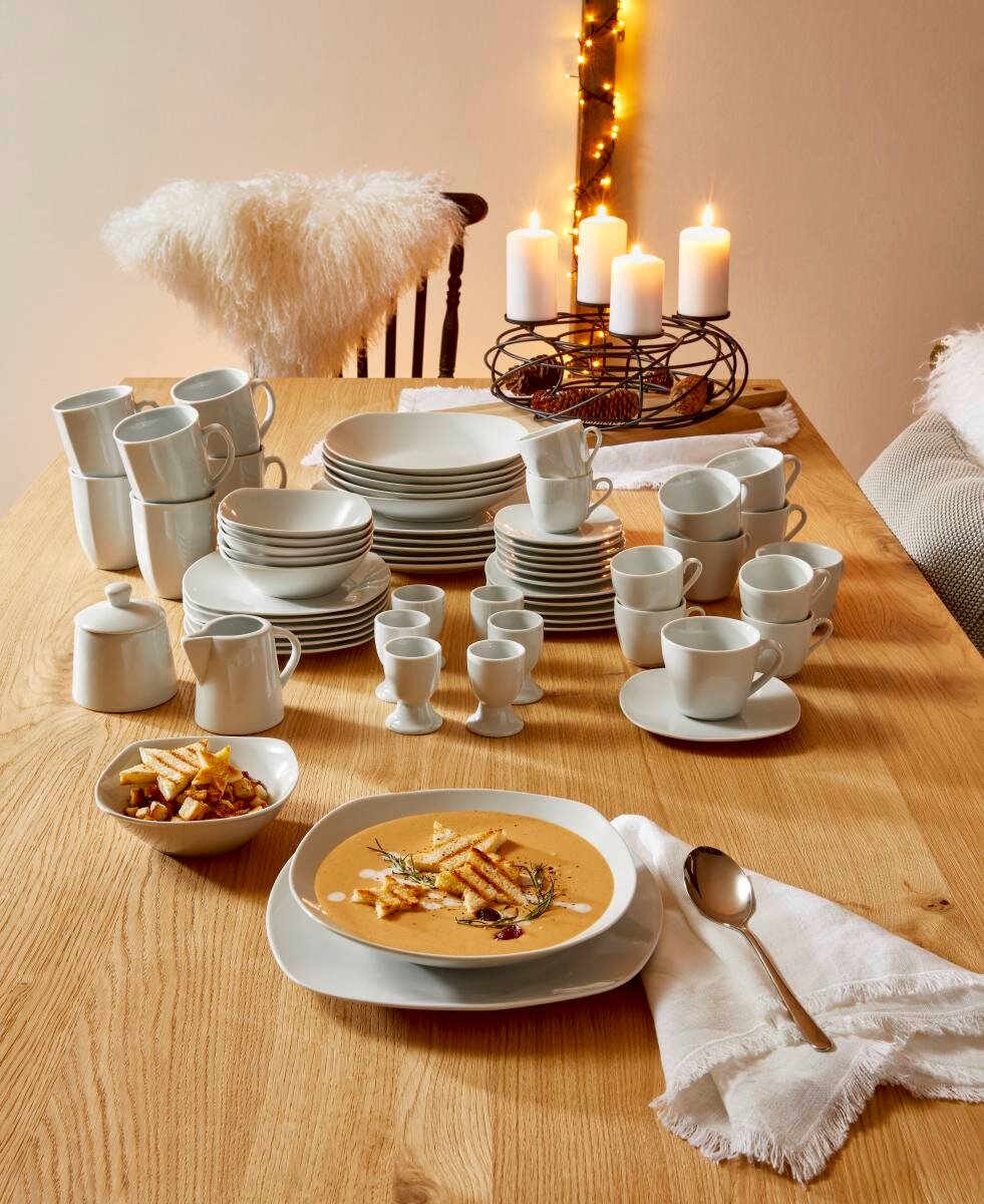 Ein 62-teiliges Kombiservice auf einem Holztisch, bestehend aus Tellern, Tassen und Schalen, mit einer gemütlichen Tischdekoration und Kerzen im Hintergrund. Perspektive von oben.
