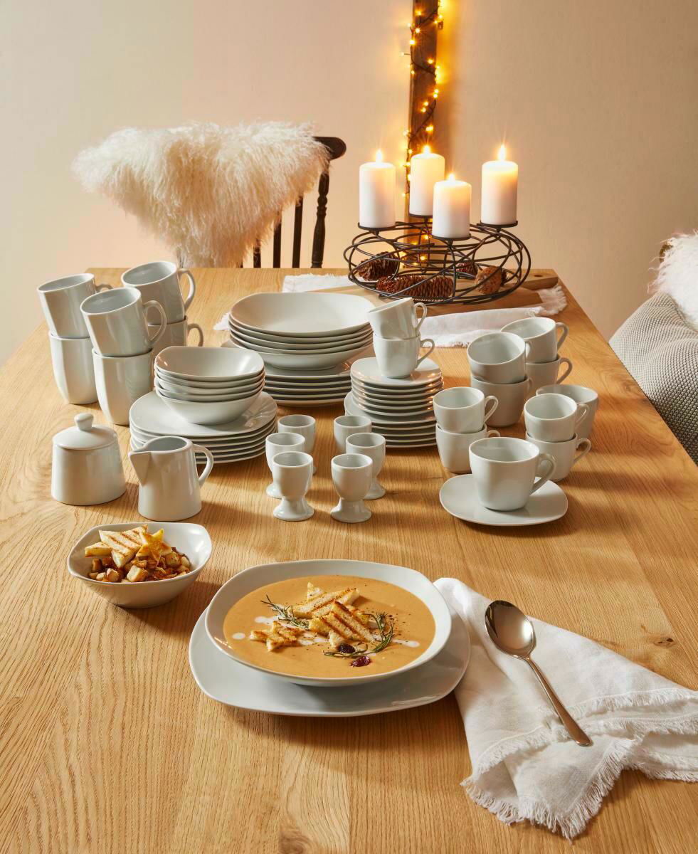 Ein 62-teiliges Kombiservice auf einem Holztisch, bestehend aus Tellern, Tassen und Schalen, mit einer gemütlichen Tischdekoration und Kerzen im Hintergrund. Perspektive von oben.