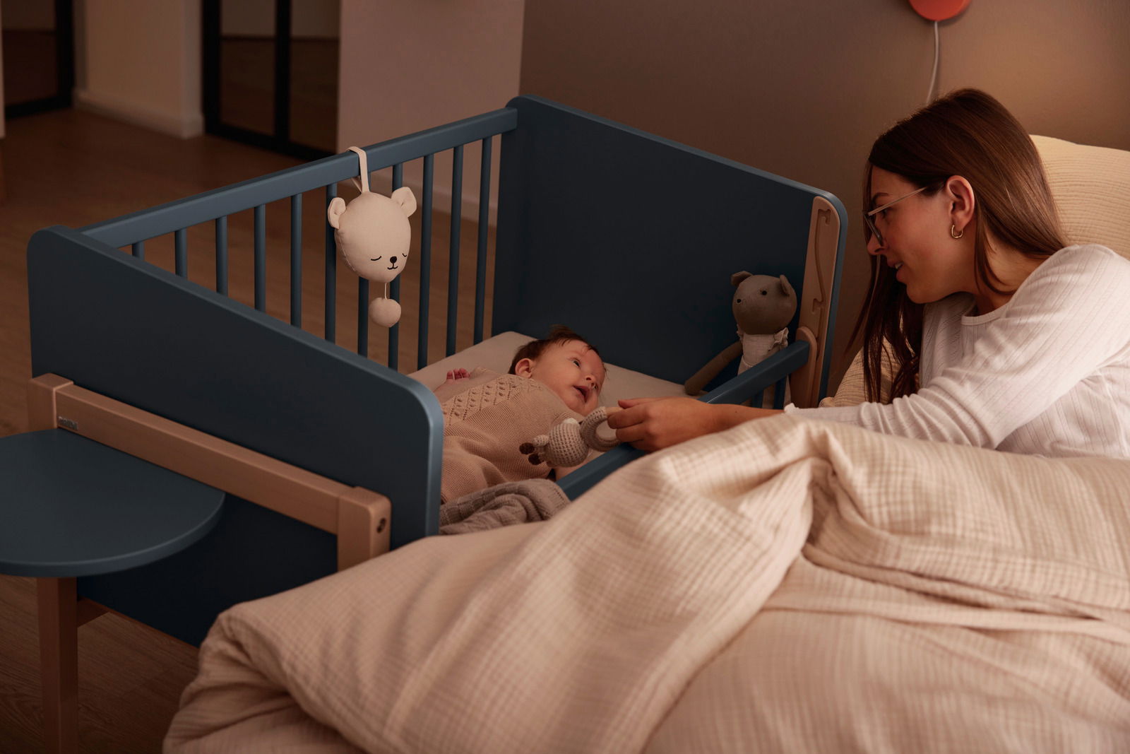 Anstellbett in einem Schlafzimmer, mit einem Baby darin, das von einer Frau liebevoll betrachtet wird. Perspektive von der Seite des Bettes.