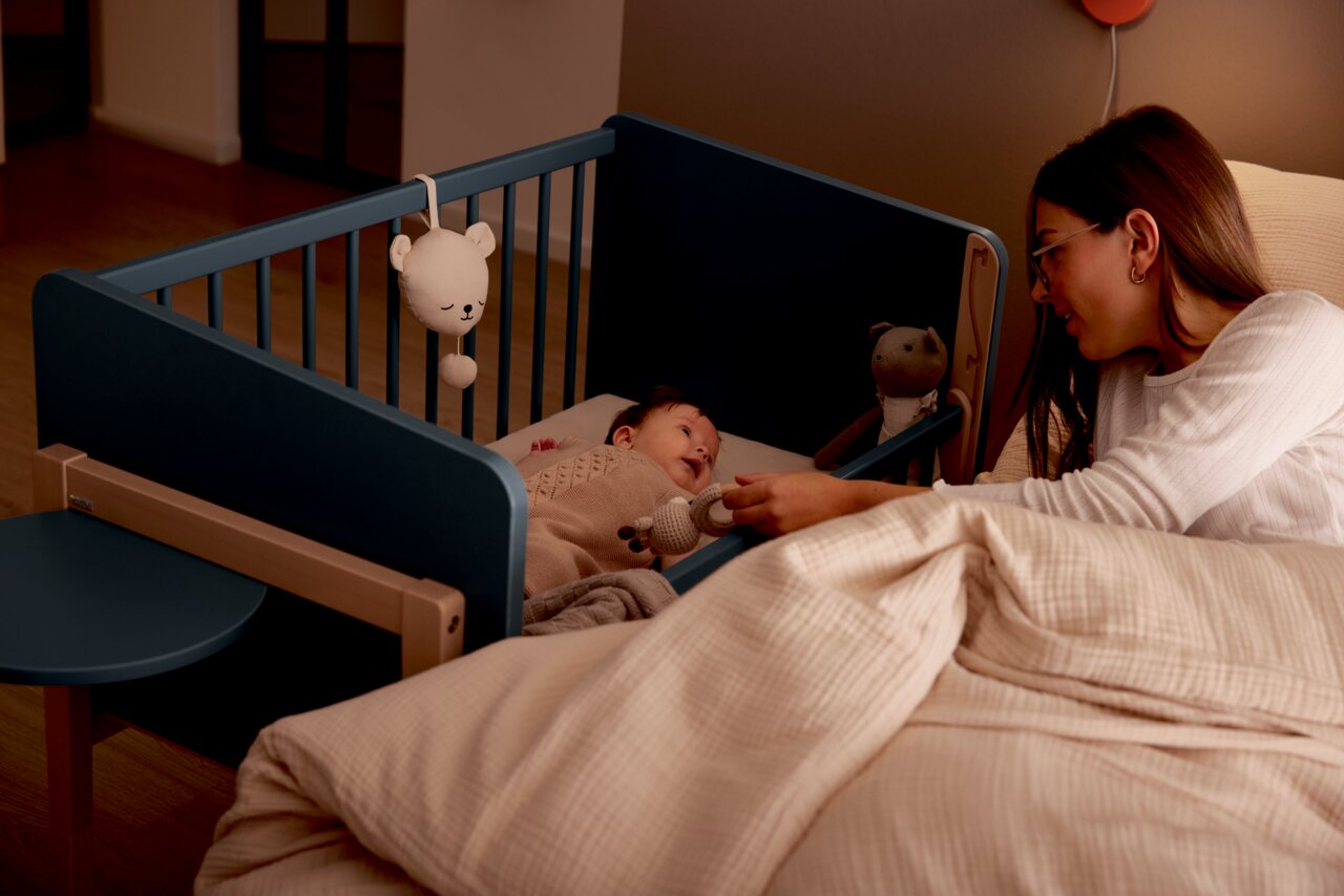 Een wieg in een slaapkamer, met daarin een baby die liefdevol door een vrouw wordt bekeken. Gezien vanaf de zijkant van het bed.