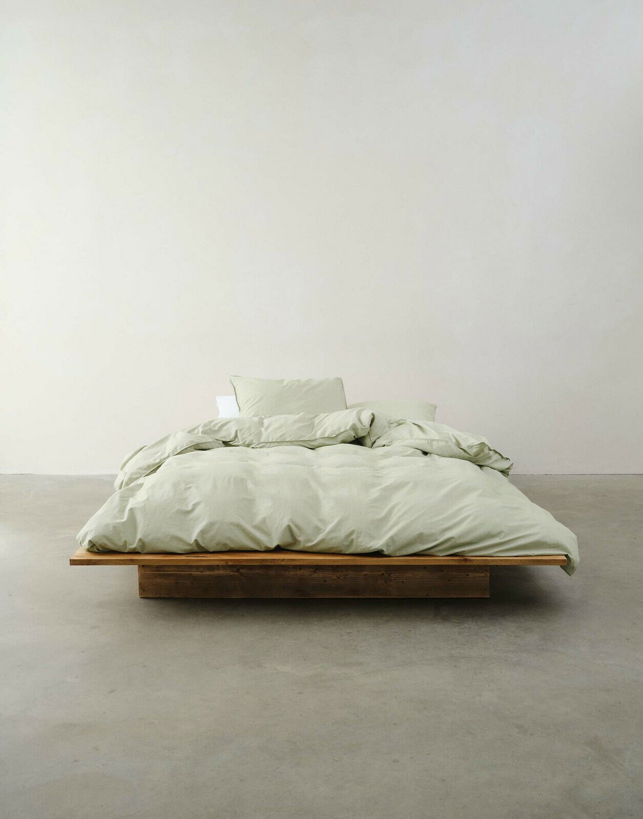 Minimalistisches Schlafzimmer mit einem niedrigen Holzbett, bezogen mit hellgrüner Bettwäsche, aus der Vorderansicht fotografiert.