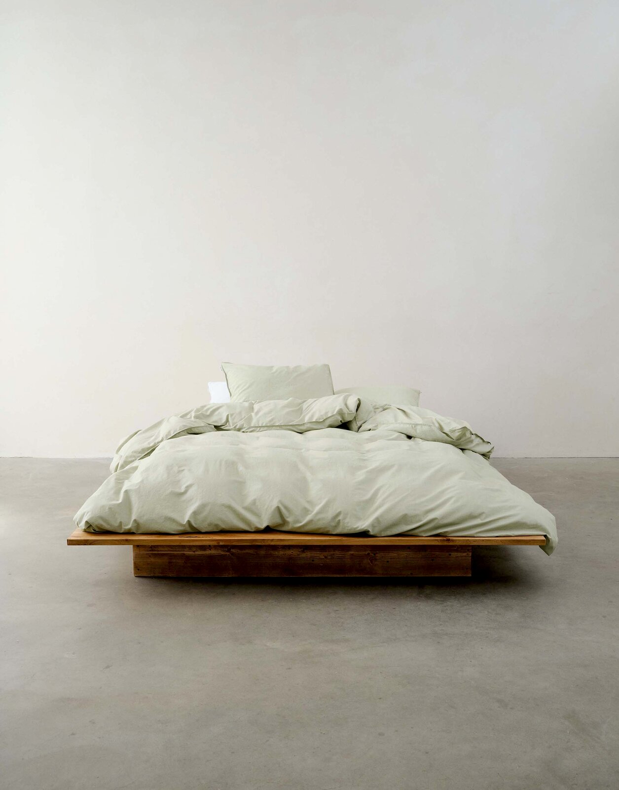 Minimalistisches Schlafzimmer mit einem niedrigen Holzbett, bezogen mit hellgrüner Bettwäsche, aus der Vorderansicht fotografiert.
