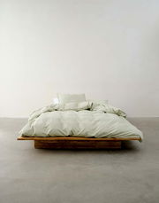 Minimalistisches Schlafzimmer mit einem niedrigen Holzbett, bezogen mit hellgrüner Bettwäsche, aus der Vorderansicht fotografiert.