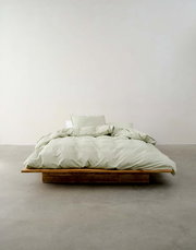 Minimalistisches Schlafzimmer mit einem niedrigen Holzbett, bezogen mit hellgrüner Bettwäsche, aus der Vorderansicht fotografiert.