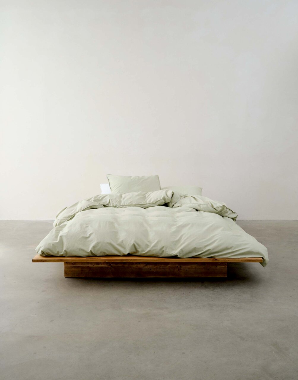Minimalistisches Schlafzimmer mit einem niedrigen Holzbett, bezogen mit hellgrüner Bettwäsche, aus der Vorderansicht fotografiert.