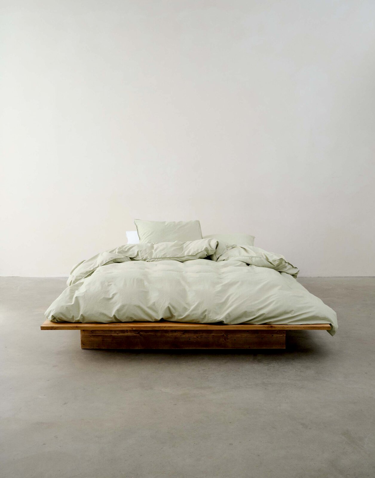 Minimalistisches Schlafzimmer mit einem niedrigen Holzbett, bezogen mit hellgrüner Bettwäsche, aus der Vorderansicht fotografiert.