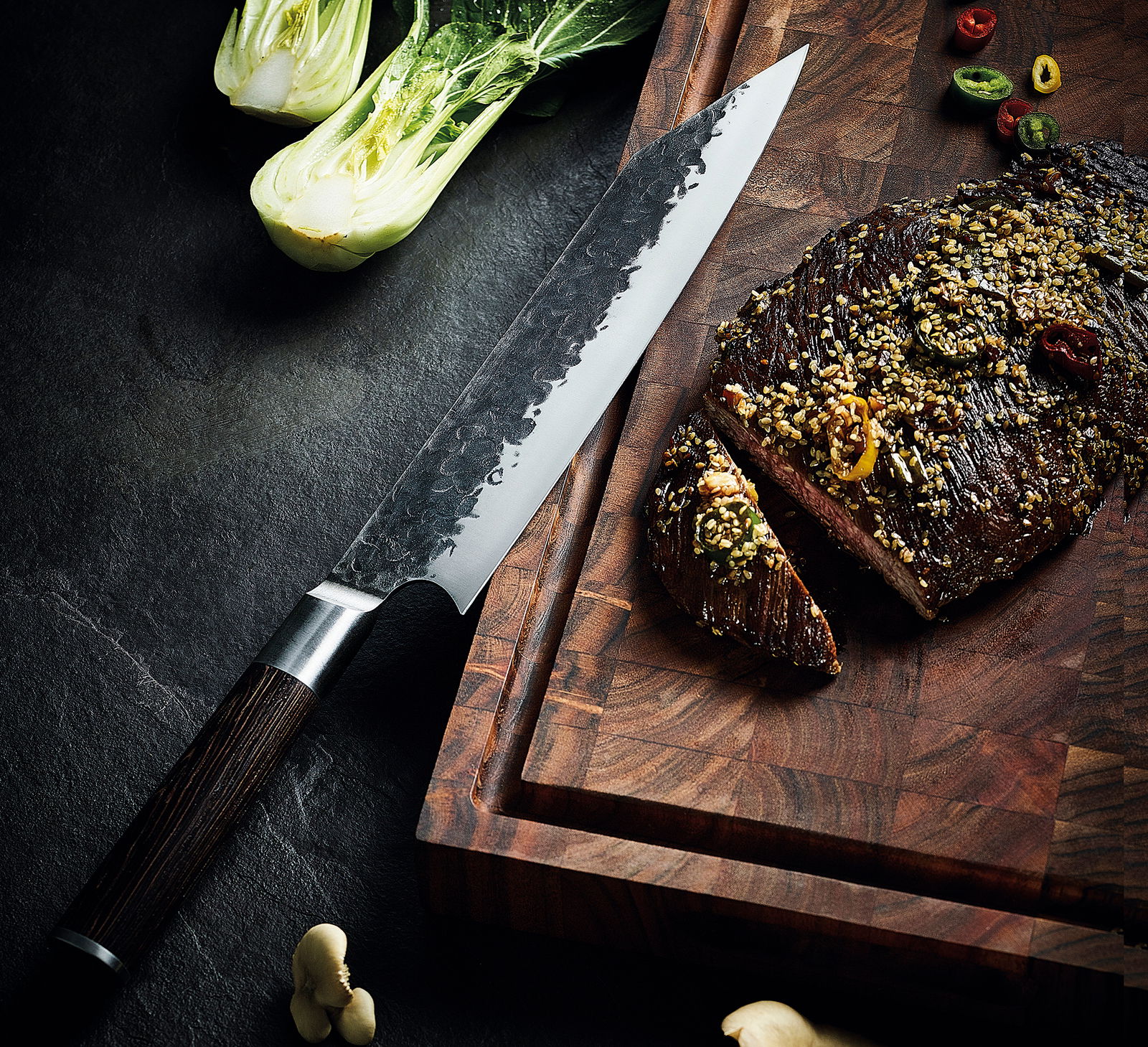Kiritsuke-Messer KYOTO 23 cm mit gehämmerter Klinge und dunklem Holzgriff, auf einem Schneidebrett neben geschnittenem Fleisch und Gemüse, aus der Vogelperspektive.