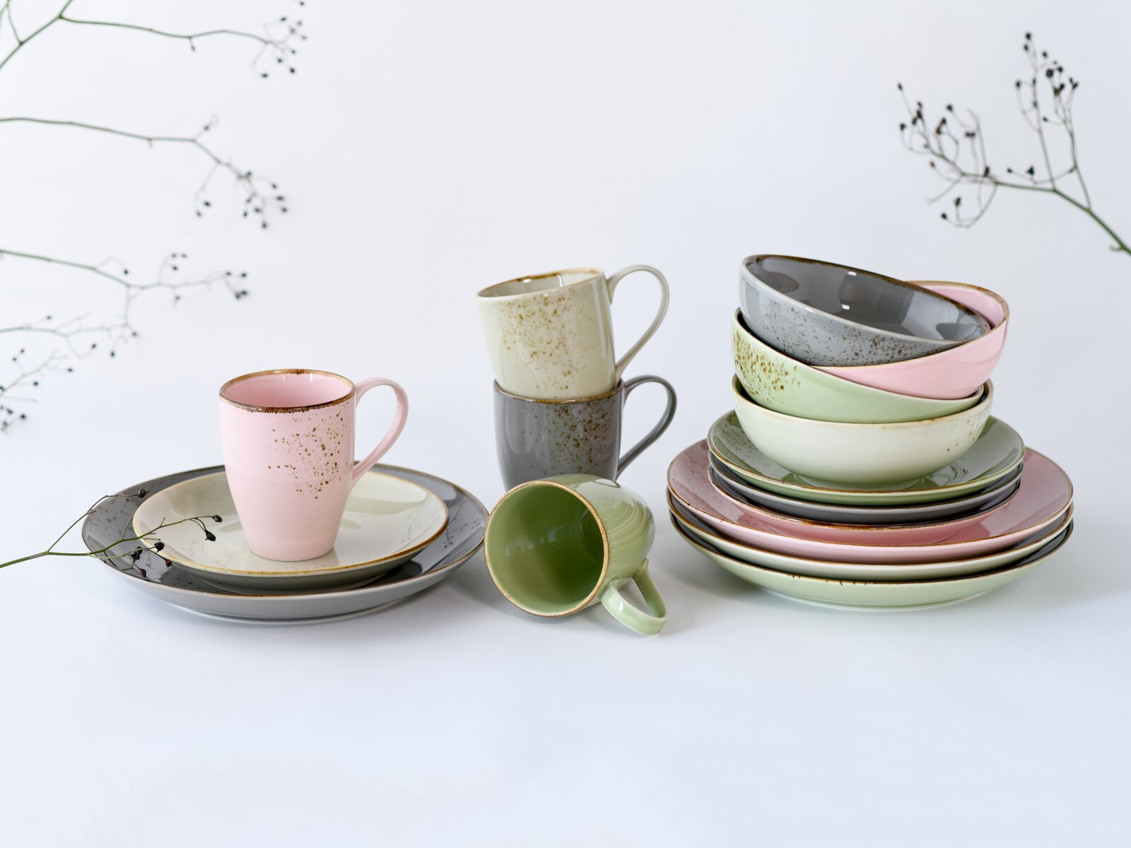 Pastellfarbenes Keramikgeschirr-Set mit Tassen, Schalen und Tellern in Rosa, Grau, Creme und Grün mit Sprenkelglasur, arrangiert auf weißem Hintergrund mit zarten Zweigen, Perspektive: leicht seitliche Frontansicht