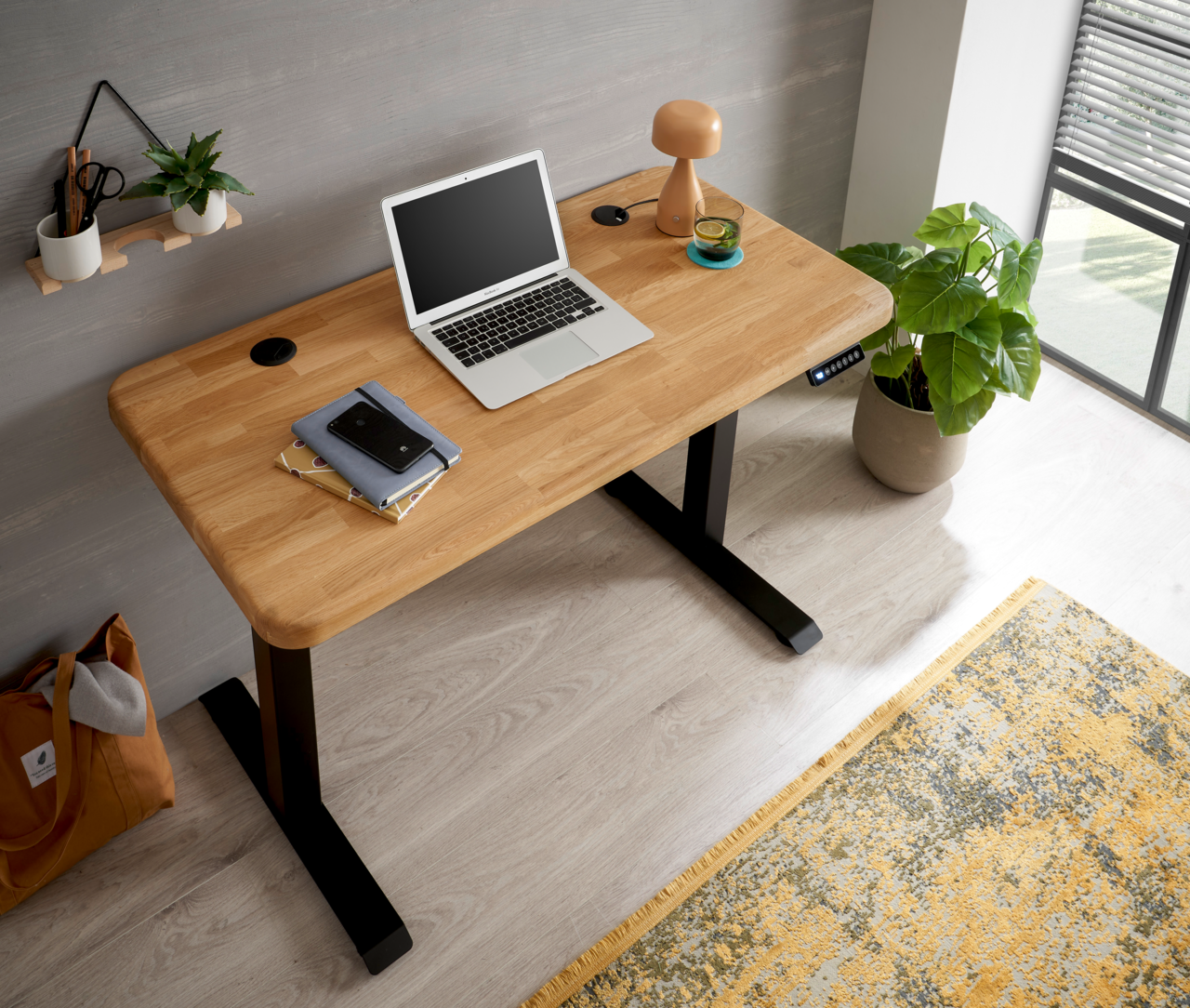 Moderner E-Schreibtisch aus Holz mit schwarzem Gestell, von oben fotografiert. Auf dem Tisch stehen ein Laptop, eine Lampe und Bücher. Eine Pflanze steht daneben.