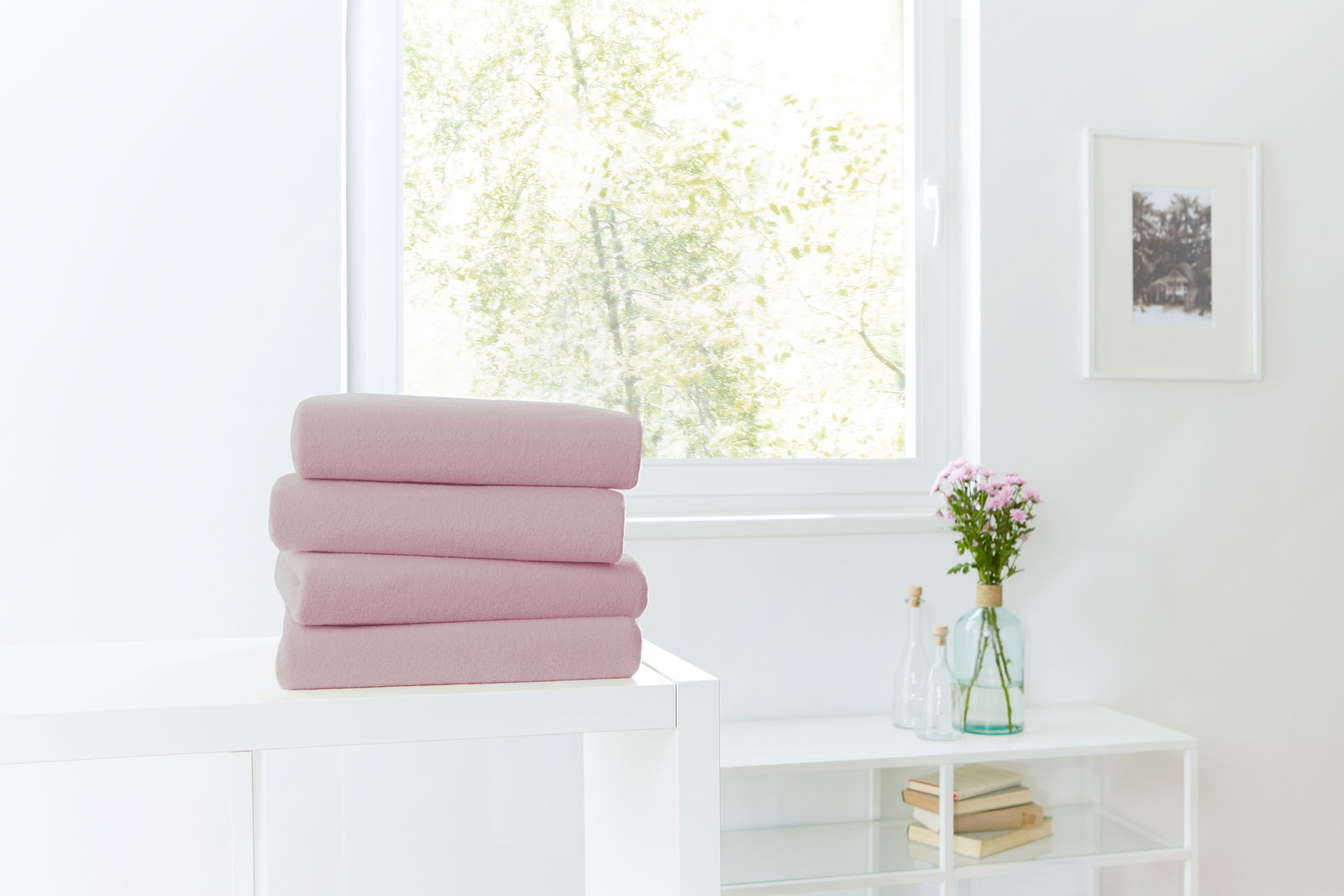 Ein Stapel von vier rosa Spannbetttüchern auf einem weißen Tisch, im Hintergrund ein Fenster mit Blick ins Grüne.