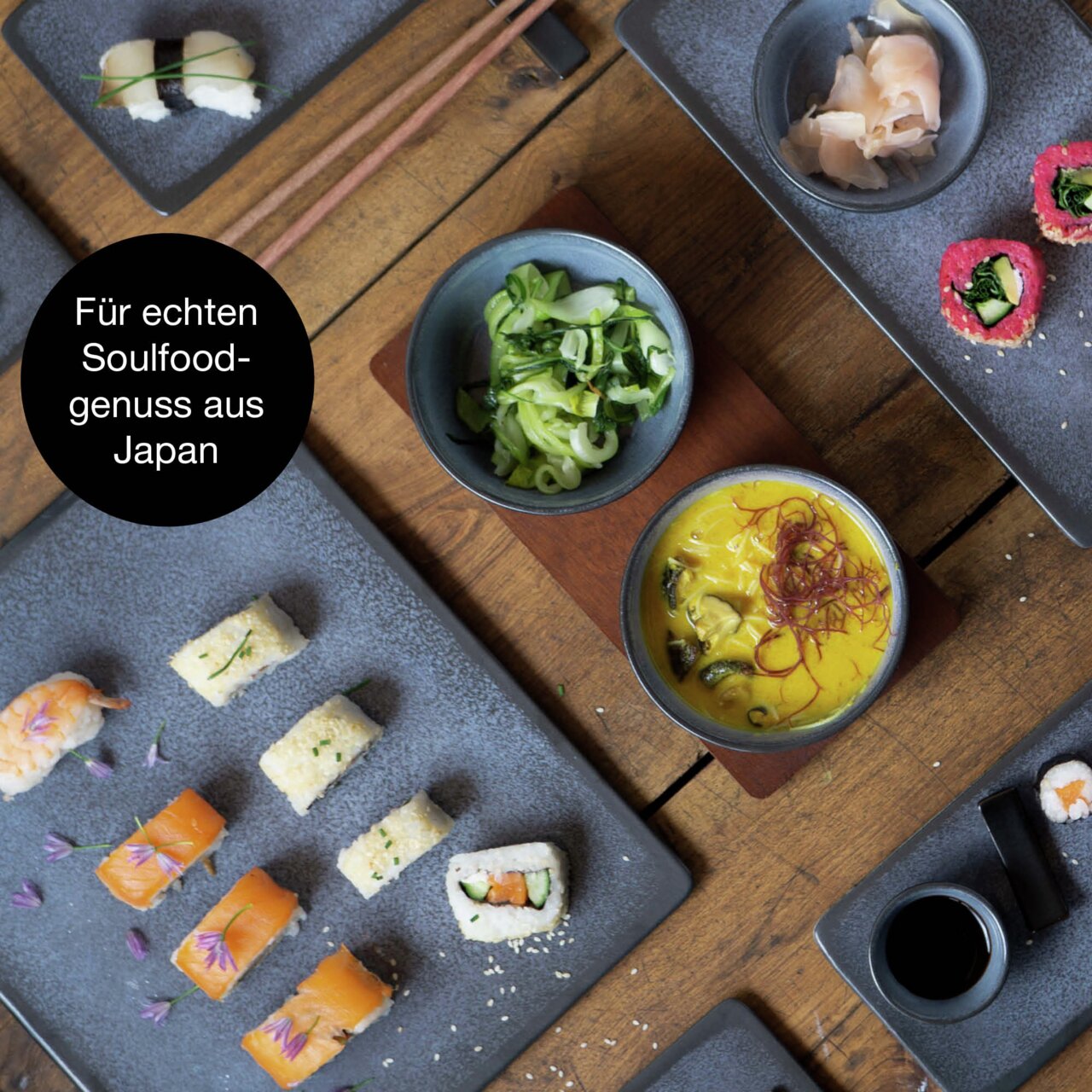 Vogelperspectief op een 29-delige sushiset met blauwe borden, gepresenteerd met verschillende soorten sushi, sojasaus en bijgerechten op een houten tafel.