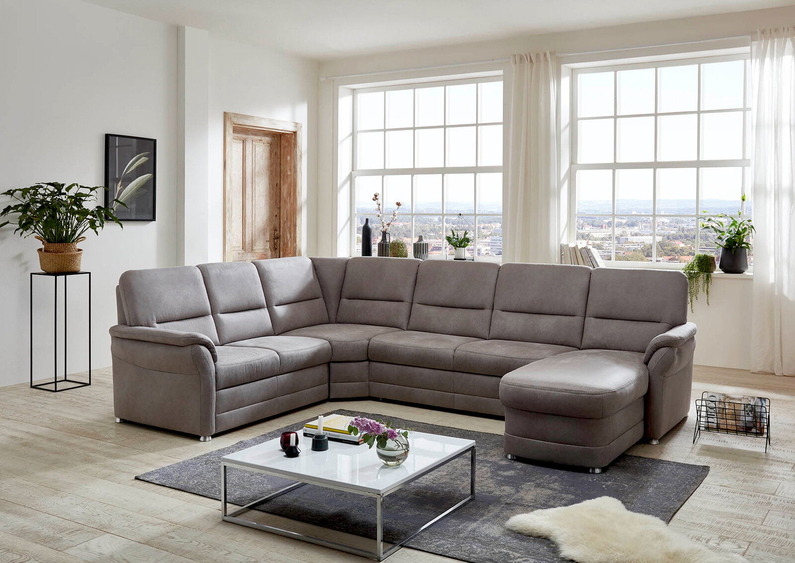 Beldomo Comfort Wohnlandschaft QUEENLINE Graue Polsterrundecke in einem modernen Wohnzimmer mit großen Fenstern, von vorne betrachtet.