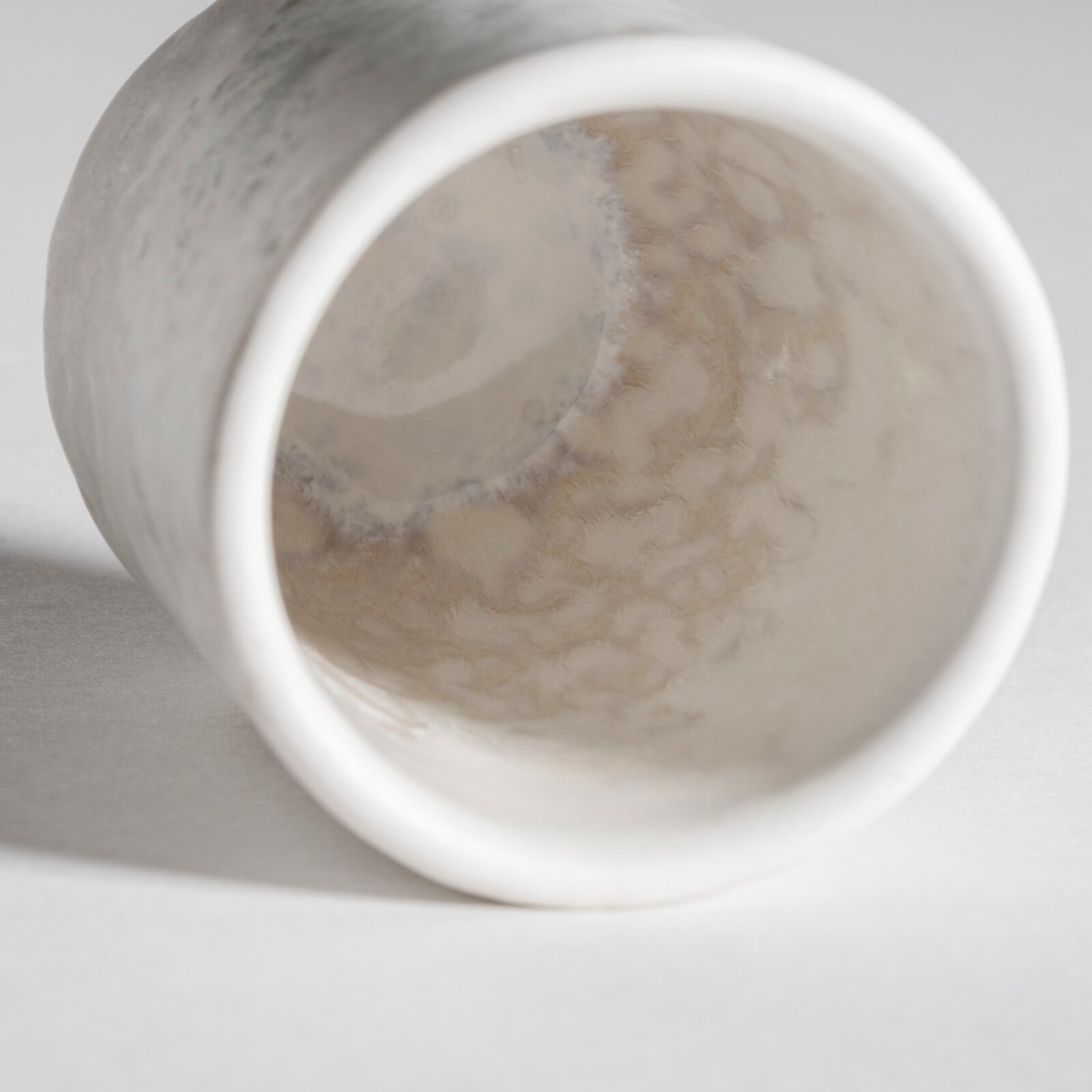 Nahaufnahme einer weiß-beigen, marmorierten Keramikvase/Becher, liegend fotografiert mit Blick in die Öffnung (Frontansicht/Top-Down-Perspektive), auf hellem Hintergrund.