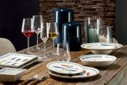 Gedeckter rustikaler Holztisch mit maritimem Fischgeschirr, eleganten Gläsern, Windlichtern und dunkelblauen Vasen in leicht schräger Vorderansicht