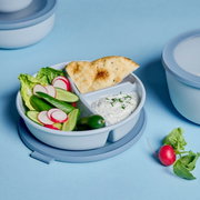 Cirqula-bento-bakje, rond en blauw, met verse groenten, flatbread en dip, gefotografeerd van bovenaf.