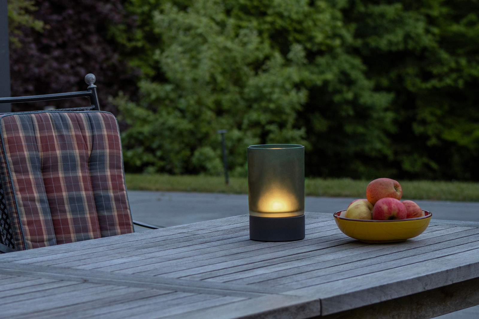 Fischer & Honsel Akku-Windlicht ARIVA PRO Akku-Windlicht auf einem Holztisch im Freien, neben einem Stuhl und einer Schale mit Äpfeln, aus seitlicher Perspektive fotografiert.