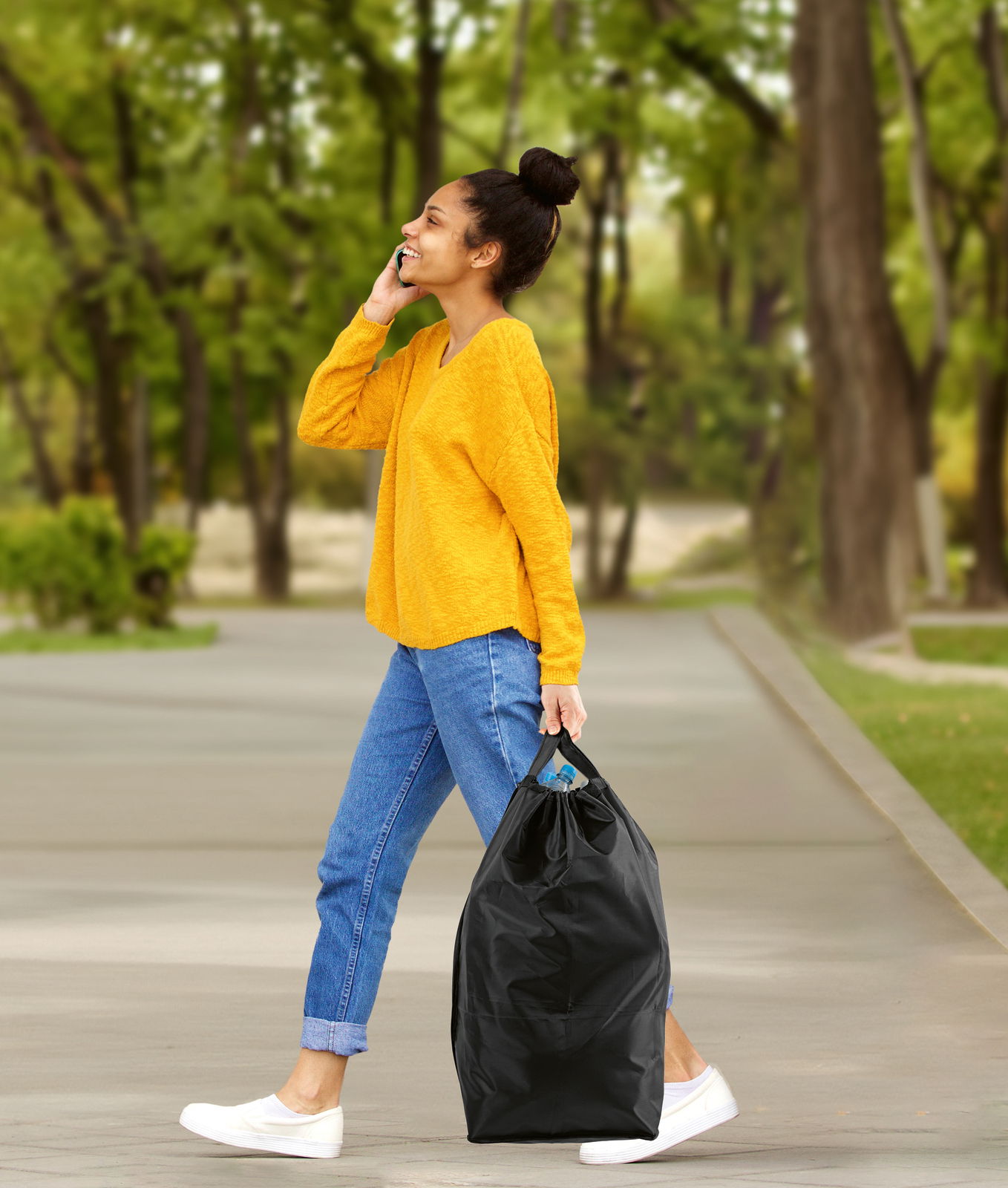 Eine Frau in gelbem Pullover und blauen Jeans geht im Park und trägt einen schwarzen Pfandflaschen Sammelbehälter in der rechten Hand. Seitenansicht.