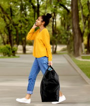 Eine Frau in gelbem Pullover und blauen Jeans geht im Park und trägt einen schwarzen Pfandflaschen Sammelbehälter in der rechten Hand. Seitenansicht.