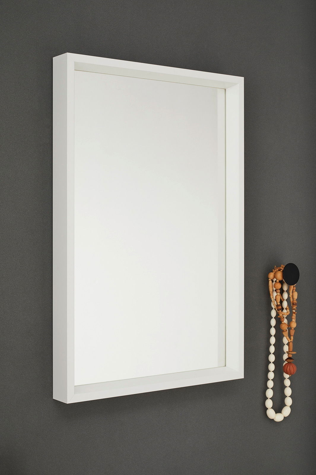 Weißer Wandspiegel mit rechteckigem Rahmen, seitliche Perspektive, an einer grauen Wand montiert, mit dekorativen Holzperlen an der Seite.