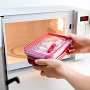 Person hält eine rote Emsa Mikrowellendose mit Einsätzen vor einer geöffneten Mikrowelle. Die Dose ist mit Lebensmitteln gefüllt und bereit zum Erhitzen. Perspektive: seitlich von vorne.