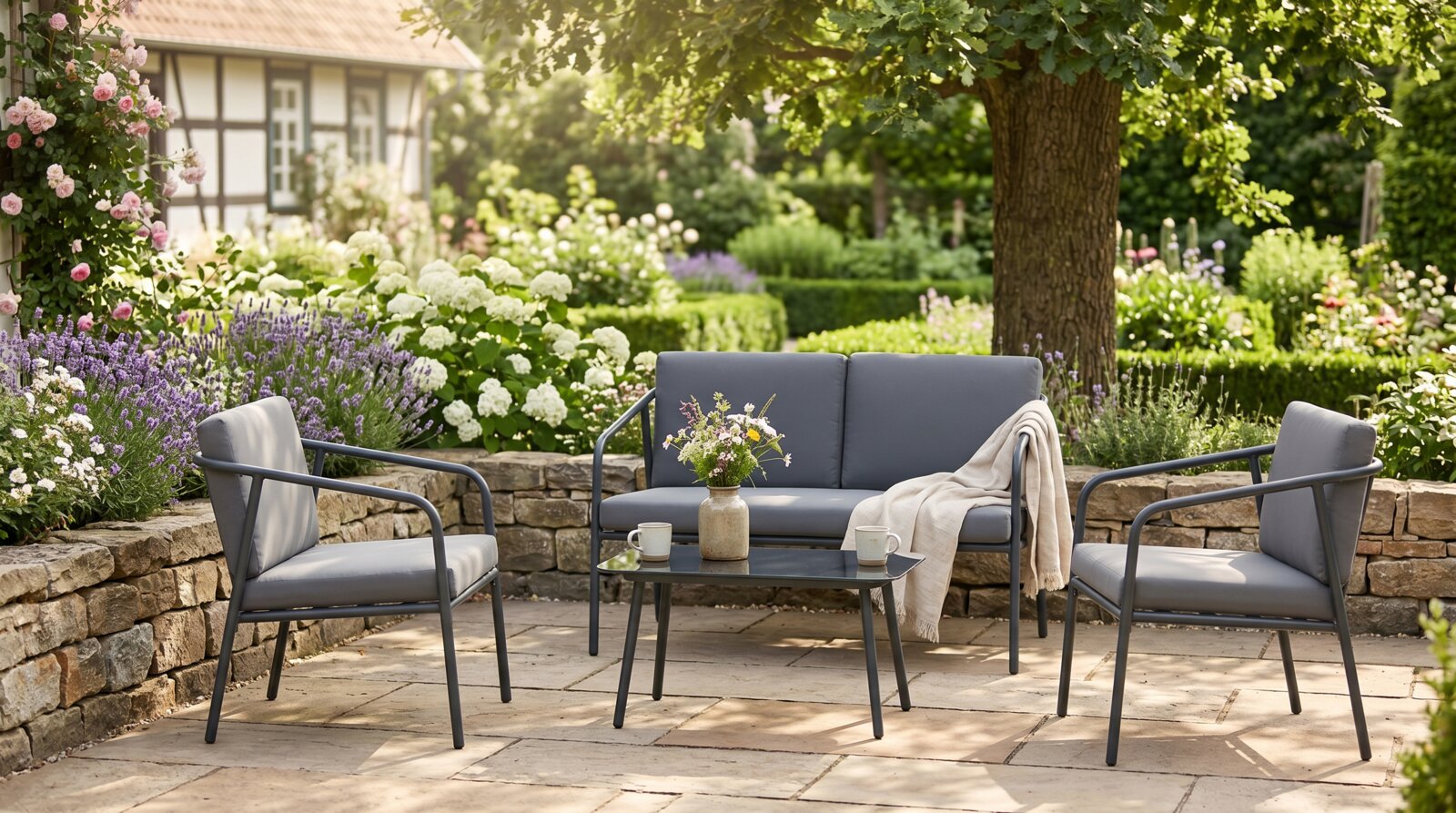 Garten-Lounge-Set in Anthrazit mit 2-Sitzer-Sofa, zwei Sesseln und niedrigem Tisch auf Stein-Terrasse, frontal aus leichter Schrägperspektive im blühenden Garten