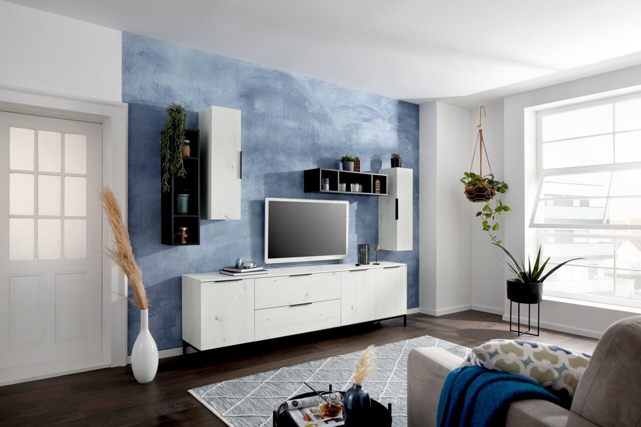 Wohnzimmeransicht mit modernem TV-Lowboard und Hängeschränken in hellem Holzdesign vor einer blauen Akzentwand, seitliche Perspektive.