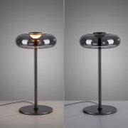 Moderne Tischleuchte mit rauchgrauem Glas-Schirm und schwarzem Metallfuß, links eingeschaltet und rechts ausgeschaltet, frontal in Augenhöhe vor grauem Hintergrund