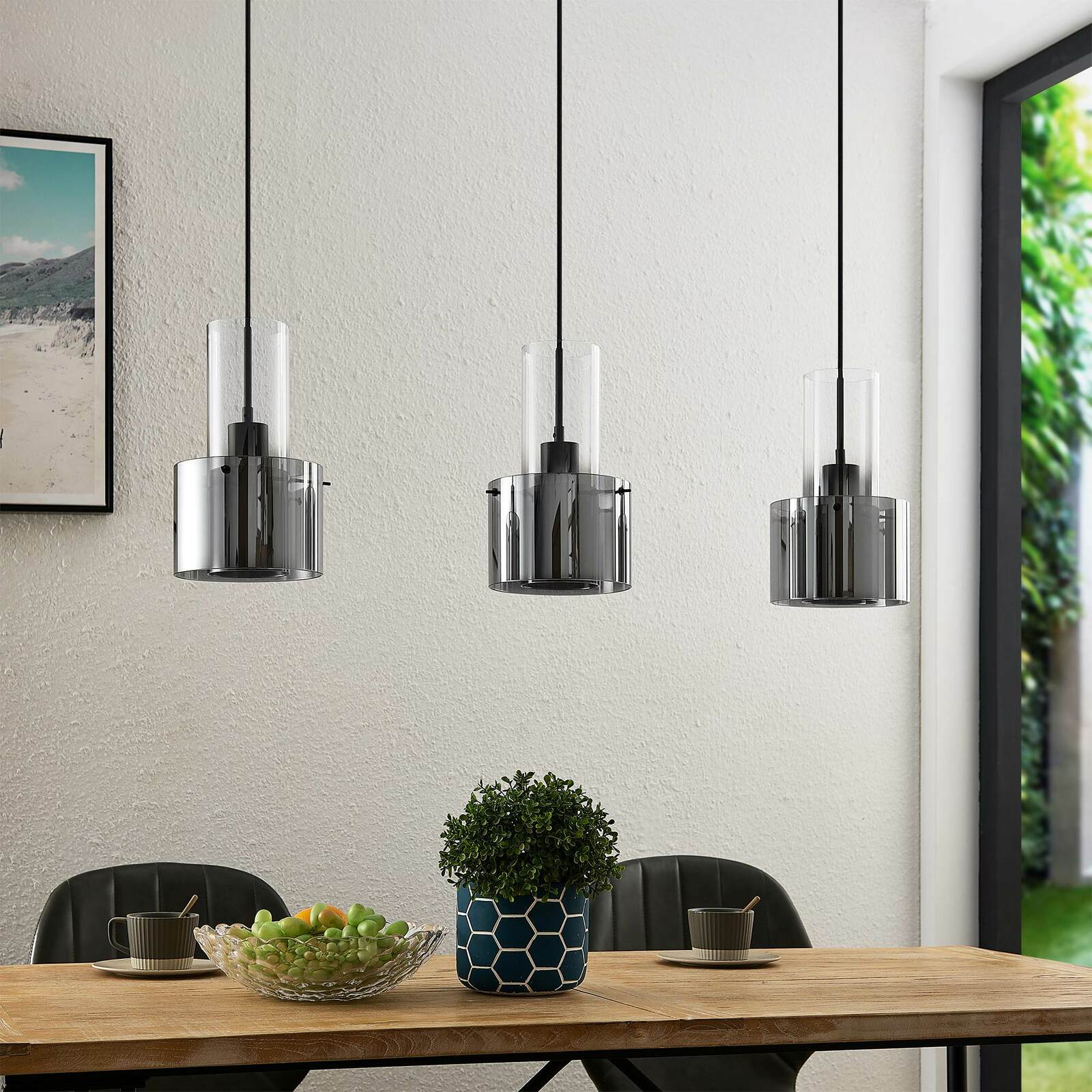Moderne Hängeleuchte mit drei zylindrischen, rauchgrauen Glasschirmen über einem Esstisch, seitliche Perspektive.