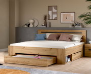 Modernes Holzbett in Eiche-Optik mit hohem Kopfteil und integrierten Schubladen/Stauraum, eine Schublade vorne ausgezogen; Ansicht schräg von vorn im Schlafzimmer-Setting.