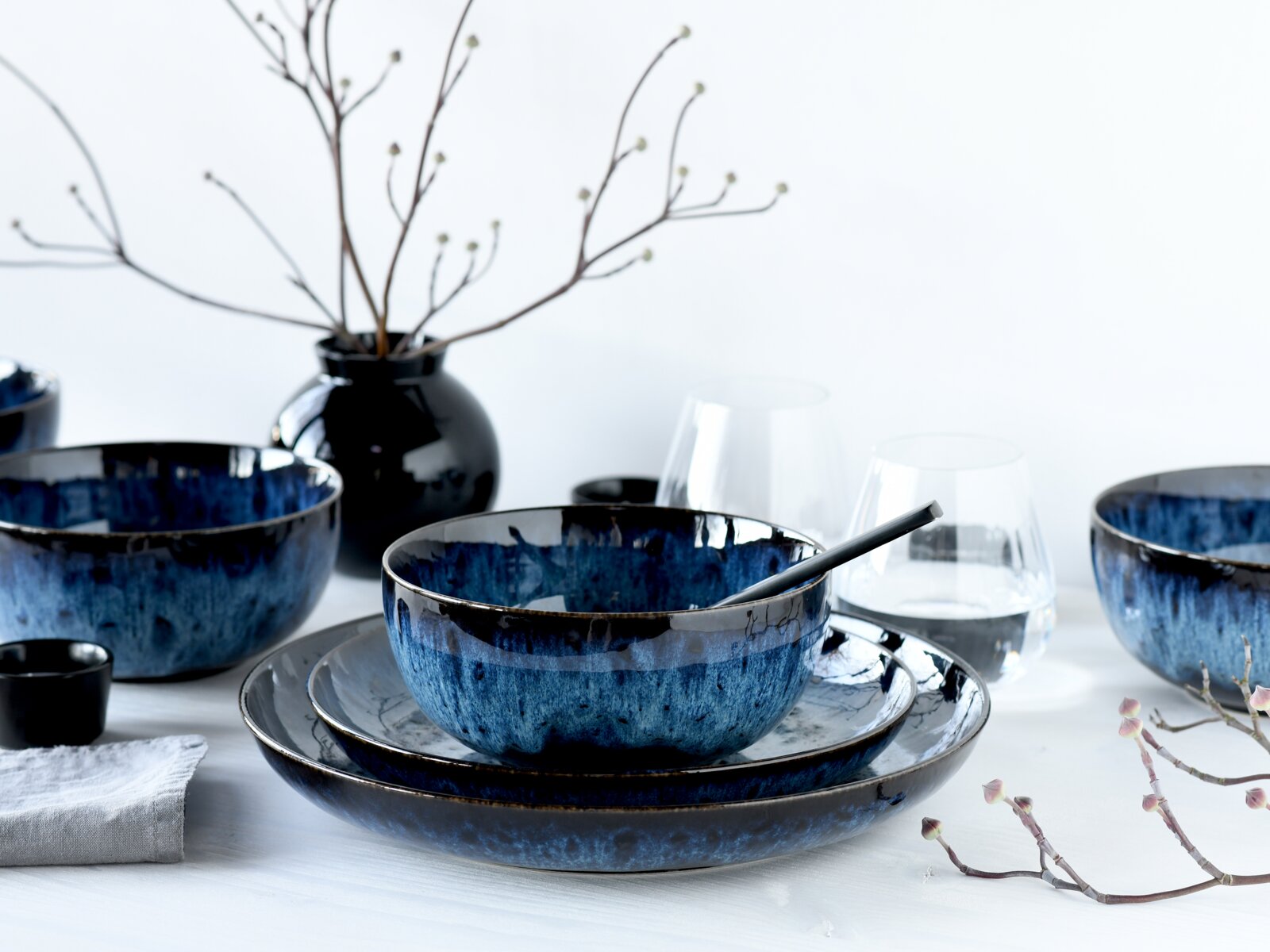 Blau-schwarz glasiertes Keramikgeschirr-Set mit Schale auf Tellern und Löffel, umgeben von weiteren Schalen und Gläsern auf weißem Tisch, Perspektive: leicht seitliche Frontansicht auf Tischhöhe