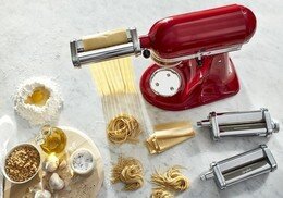 KitchenAid Nudelvorsatz (3Walzen) Edelstahl Roter Küchenmaschinenaufsatz mit Nudelvorsatz, der frische Pasta herstellt, umgeben von Zutaten und weiteren Walzenaufsätzen, aus der Vogelperspektive.