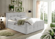 Boxspringbett in einem hellen Schlafzimmer mit Pflanzen und großen Fenstern, seitliche Perspektive