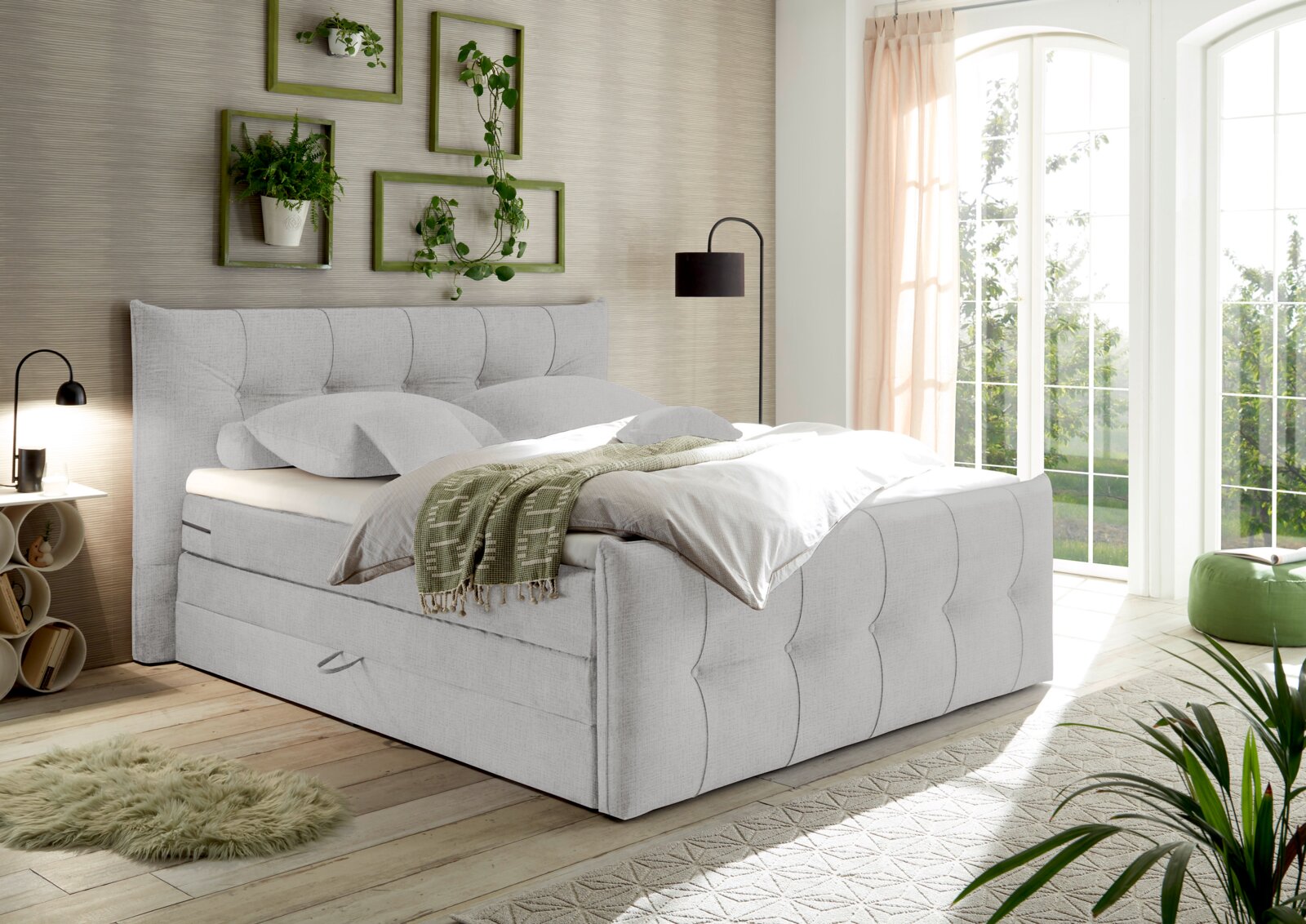 Boxspringbett in einem hellen Schlafzimmer mit Pflanzen und großen Fenstern, seitliche Perspektive