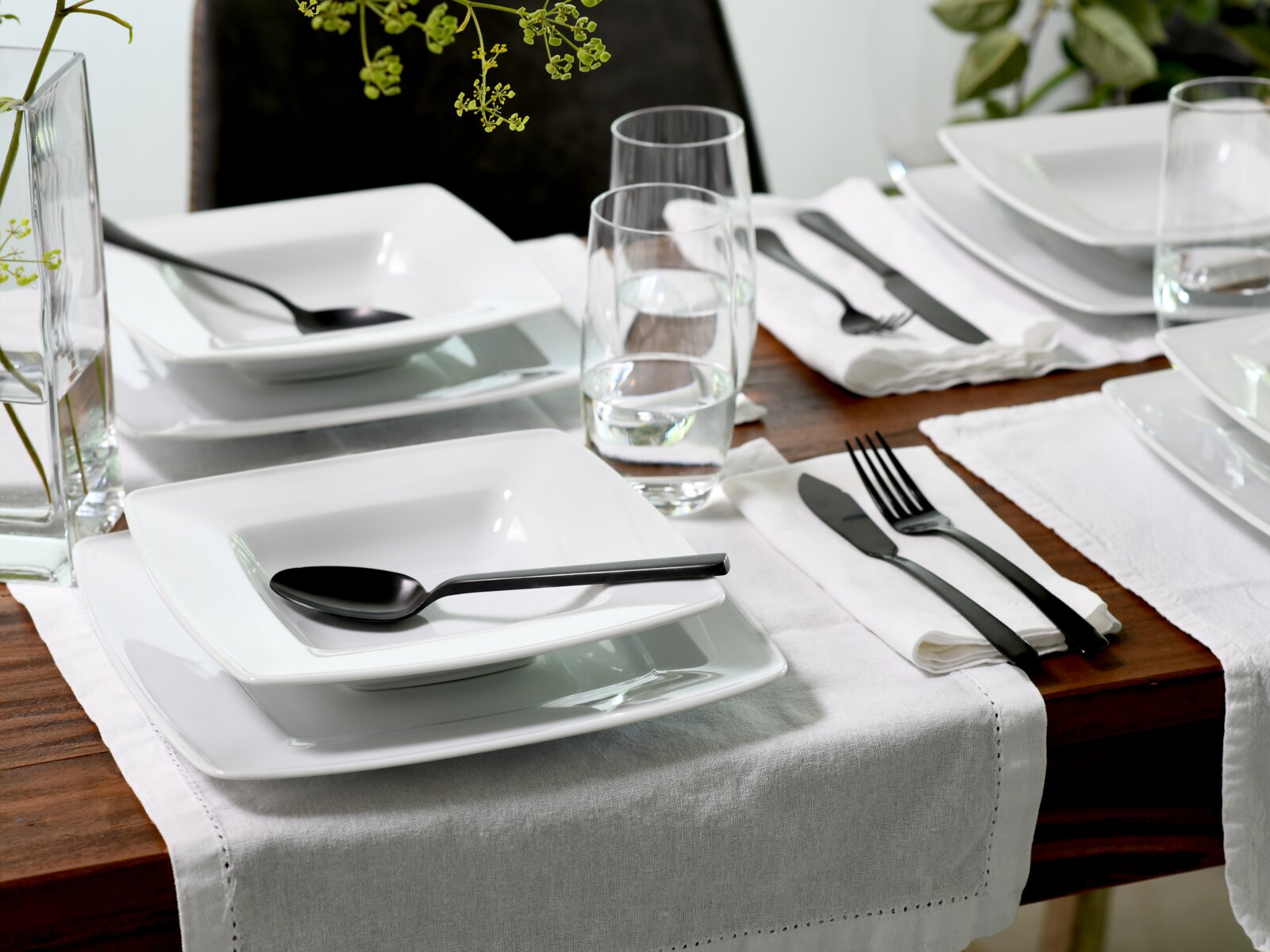 Modern gedeckter Esstisch mit weißen, quadratischen Tellern und Schalen, schwarzen Besteckteilen, Wassergläsern und weißem Tischläufer auf Holztisch, schräg von oben fotografiert.