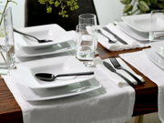 Modern gedeckter Esstisch mit weißen, quadratischen Tellern und Schalen, schwarzen Besteckteilen, Wassergläsern und weißem Tischläufer auf Holztisch, schräg von oben fotografiert.