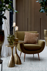 Moderner Beistelltisch mit goldener Oberfläche, dekoriert mit einer grünen Vase und Kerzen, neben einem olivgrünen Sessel. Perspektive: seitlich.