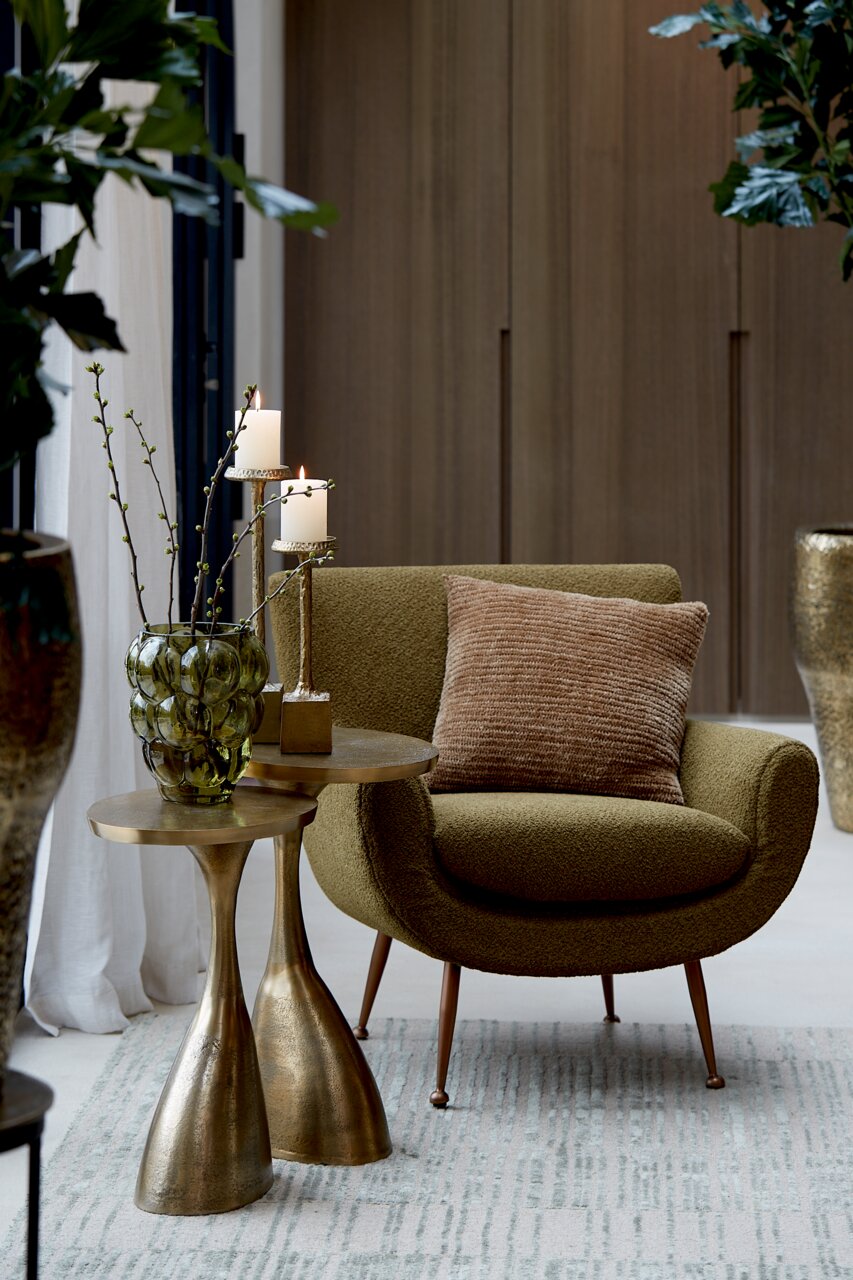 Moderner Beistelltisch mit goldener Oberfläche, dekoriert mit einer grünen Vase und Kerzen, neben einem olivgrünen Sessel. Perspektive: seitlich.