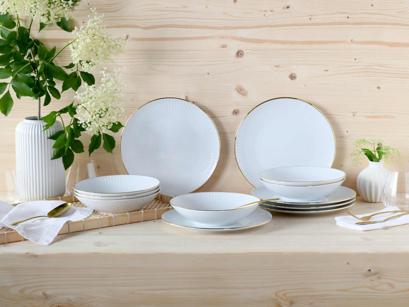 Creatable Tafelservice 12-tlg VALENCIA Frontale Tischansicht eines eleganten weißen Porzellan-Geschirrsets mit feiner Rillenstruktur und goldfarbenem Rand, arrangiert auf hellem Holztisch mit Vasen, Blumen und goldfarbenem Besteck.