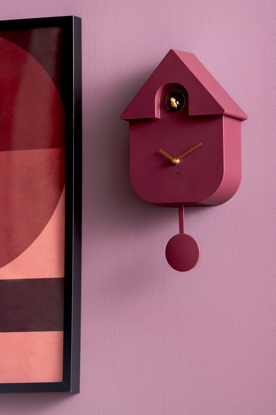 Moderne roze wandklok in de vorm van een koekoeksklok met gouden wijzers en slinger, gezien vanaf de zijkant.