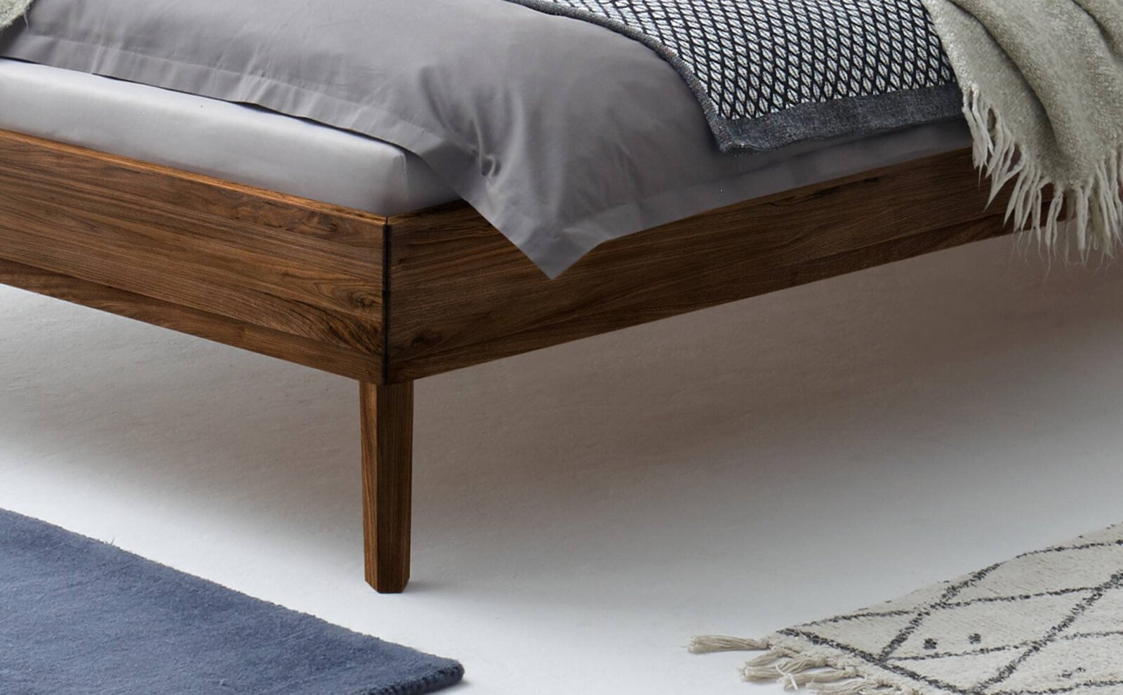 Modern houten bedframe met zichtbare houtnerf en conische poten, grijs beddengoed en deken, detailopname vanuit een schuin zijaanzicht op de hoek van het bed