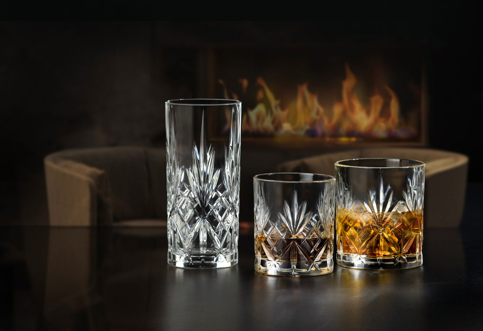 Drei Kristallgläser in verschiedenen Größen, elegant vor einem Kamin arrangiert, mit einem gemütlichen Sessel im Hintergrund.