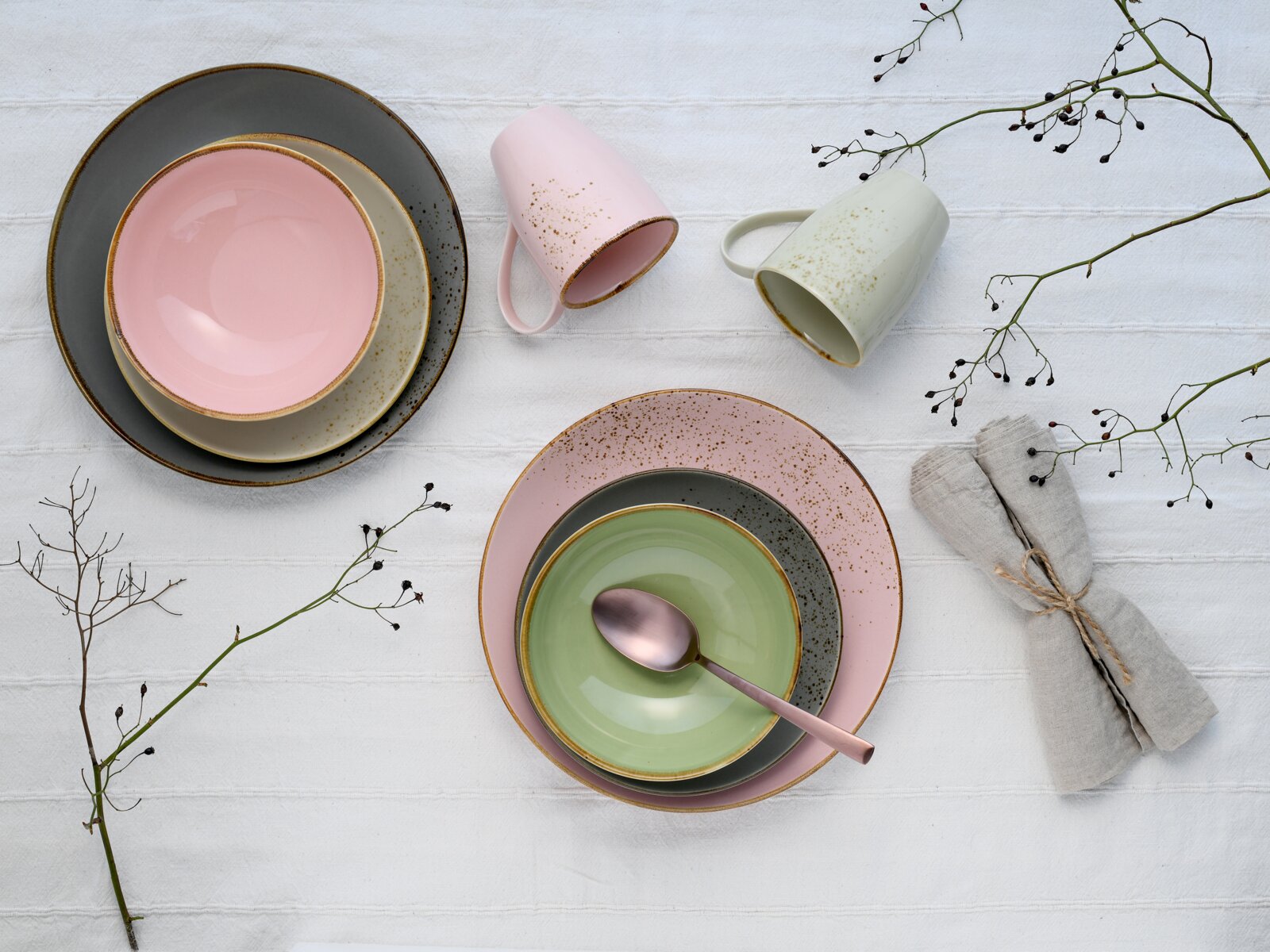 Pastellfarbenes Keramik-Geschirrset mit Tellern und Schalen in Rosa, Grau und Grün sowie zwei Tassen und Löffel auf weißer Tischdecke, Draufsicht (von oben)