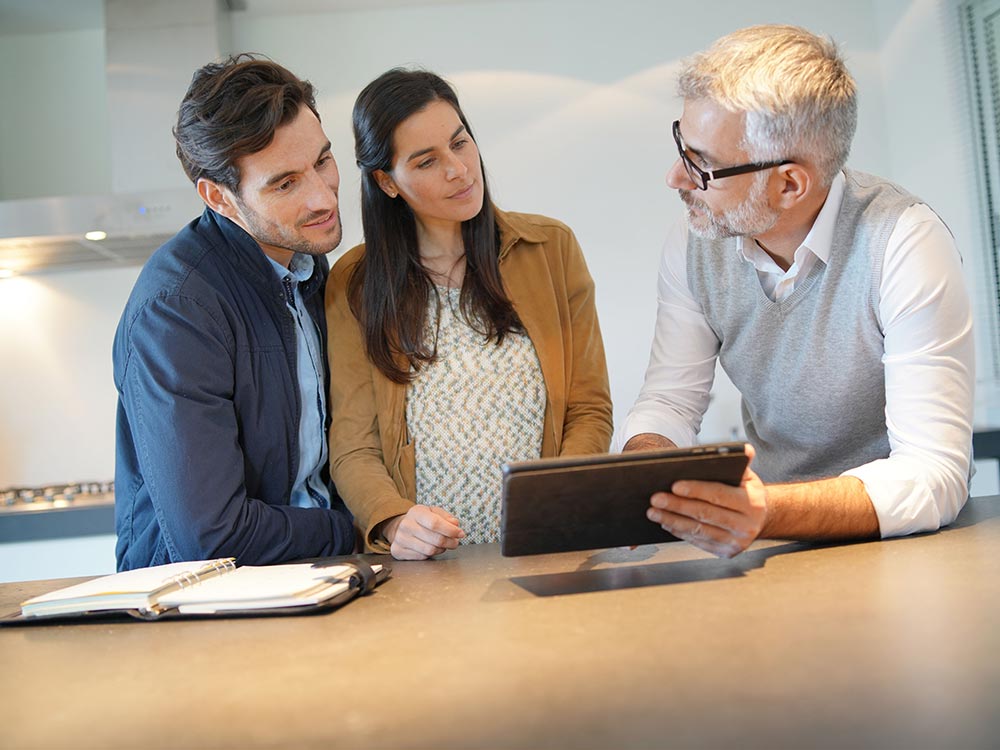 Ein älterer Mann erklärt einem jungen Paar etwas auf einem Tablet. Der Mann trägt eine weiße Bluse mit grauer Weste, der junge Mann eine dunkelblaue Jacke und die Frau eine beige Jacke mit gemustertem Oberteil. Sie stehen an einer modernen Kücheninsel mit