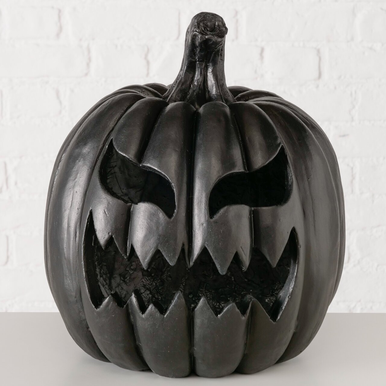 Schwarzer Halloween-Kürbis (Jack-o’-Lantern) mit grimmigem, ausgeschnittenem Gesicht und gezacktem Mund, frontal aus leicht erhöhter Perspektive vor weißer Ziegelwand