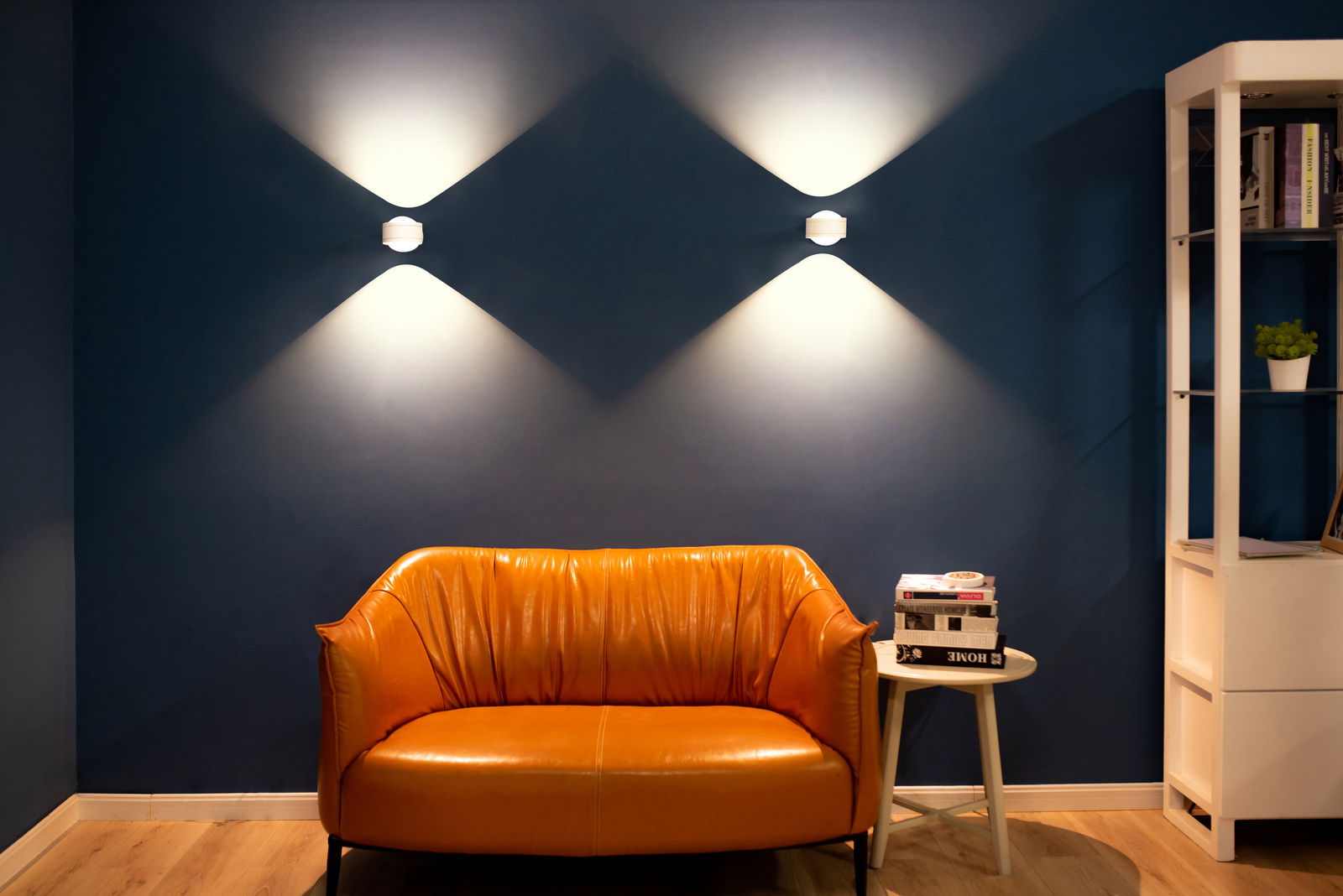 Zwei LED Wandleuchten an einer blauen Wand, die ein stilvolles Lichtmuster erzeugen, mit einem orangefarbenen Sofa im Vordergrund. Perspektive: Frontalansicht.