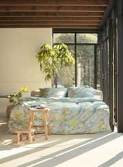 Gemütliches Schlafzimmer mit Bettwäsche in floralen Mustern, Tageslicht durch große Fenster, Pflanzen im Hintergrund, seitliche Perspektive.