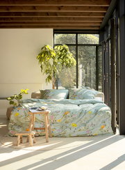 Gemütliches Schlafzimmer mit Bettwäsche in floralen Mustern, Tageslicht durch große Fenster, Pflanzen im Hintergrund, seitliche Perspektive.