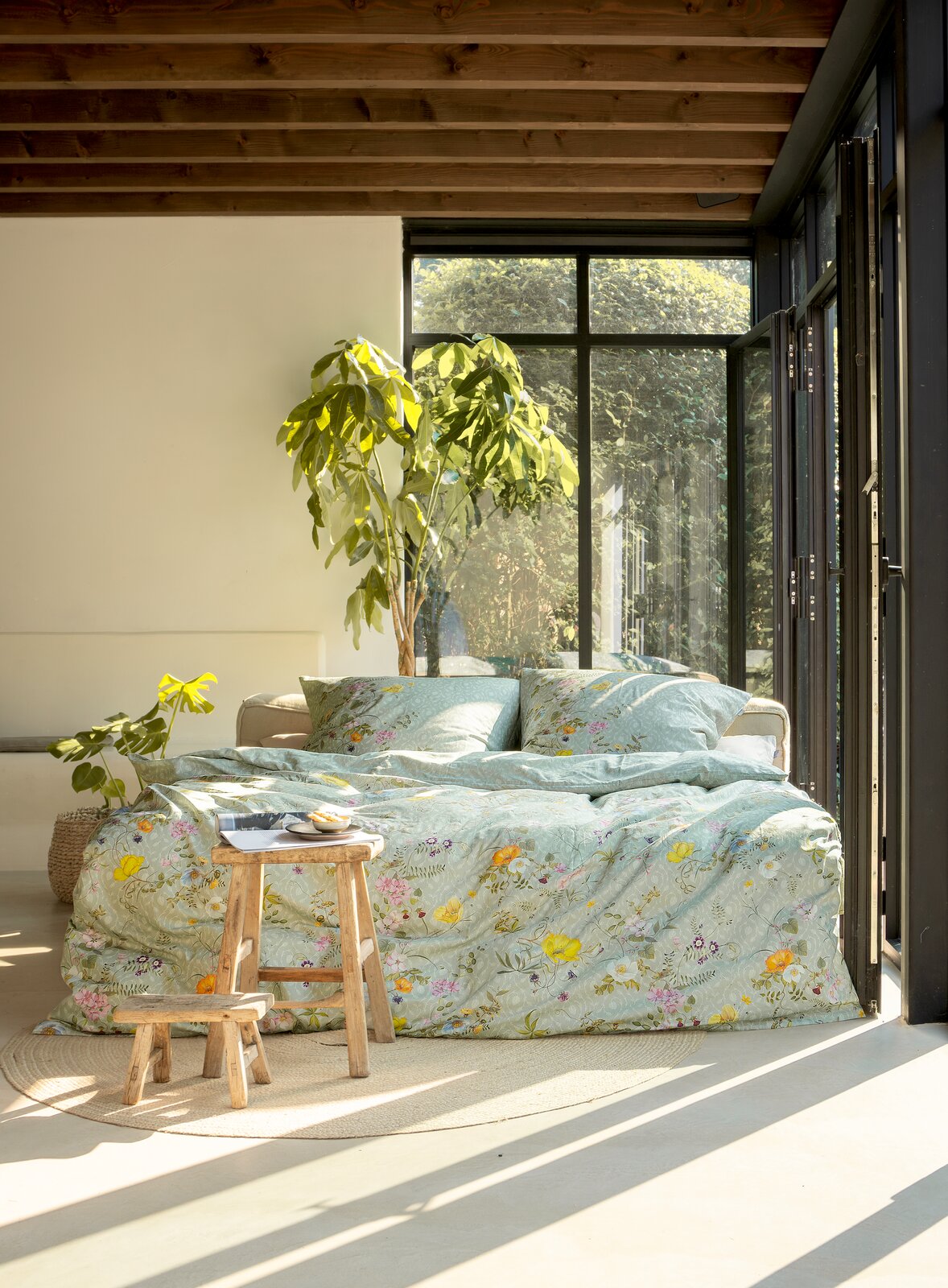 Gemütliches Schlafzimmer mit Bettwäsche in floralen Mustern, Tageslicht durch große Fenster, Pflanzen im Hintergrund, seitliche Perspektive.