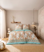 Bettwäsche-Set in einem gemütlichen Schlafzimmer, mit Paisley-Muster in Orange und Grün auf weißem Hintergrund, aus der Perspektive von vorne aufgenommen.